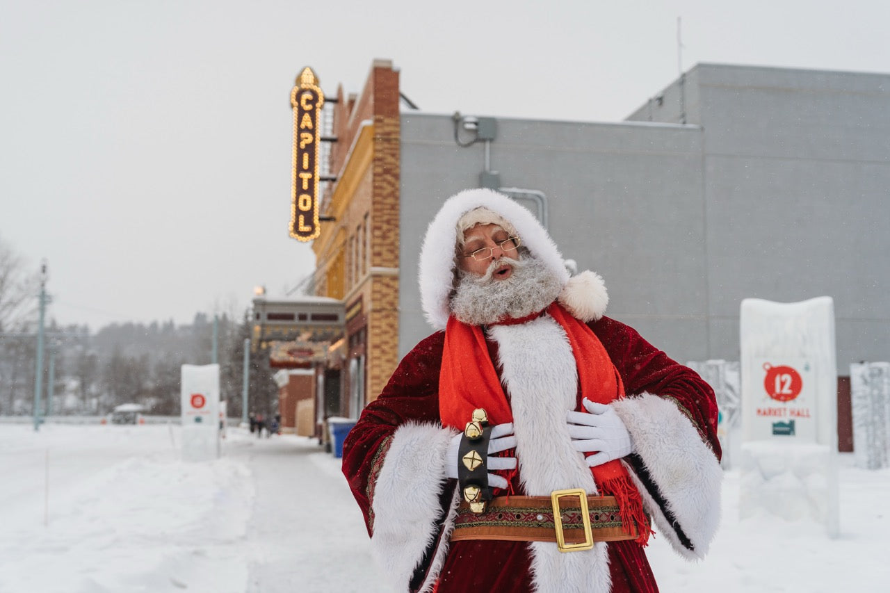 Man dressed as Santa Claus standing in a snowy landscape with a building and sign in the background.